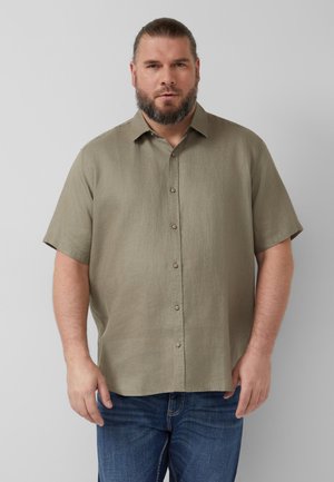 Man met korte baard, die een beige kortemouw blouse met knopen en blauwe spijkerbroek draagt, staand tegen een lichtgrijze achtergrond.