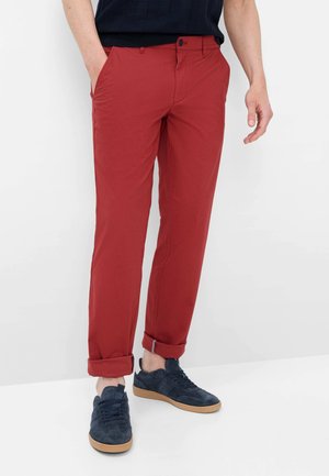 Mann trägt hochgekrempelte rote Hose, schwarzes Hemd und dunkelblaue Sneakers mit Gummisohlen, steht mit einer Hand in der Tasche.