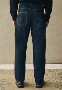 Jeans de mezclilla de un azul oscuro con corte recto, que cuentan con bolsillos traseros cosidos y detalles en hilo dorado, combinados con calzado negro.