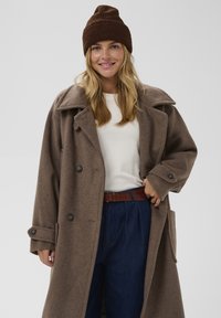 Palton dintr-un amestec de lână de culoare maro, cu revere mari, nasturi dubli și buzunare laterale, purtat peste un pulover alb și pantaloni albaștri. Beanie maro.