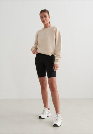 Sweatshirt beige avec un col rond et des détails textuels, associé à un short de cyclisme noir et des baskets blanches sur un fond neutre.