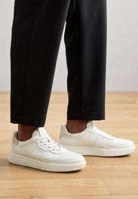 Vita lädersneakers med perforerad detalj, platta snören och en texturerad ovandel. Rundad tå och tjock sula. Kombinerat med svarta byxor.