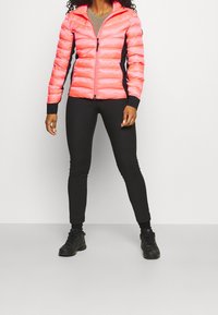 Kobieta stoi w jasnoróżowej kurtce puchowej, czarnych legginsach i czarnych butach sportowych na jednolitym jasnym tle.