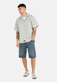 Camisa de botones de manga corta en verde claro con un corte relajado, combinada con pantalones cortos de mezclilla azul. La vestimenta presenta un estilo casual y desenfadado.