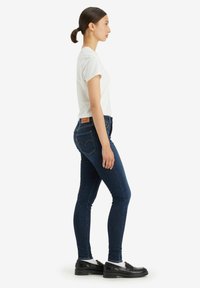 Jeans skinny de denim azul oscuro con un parche trasero de marca, combinados con una camiseta blanca ajustada y mocasines negros, vista lateral sobre un fondo blanco.