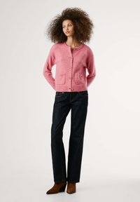 Model poseert met handen op de heupen, gekleed in een roze cardigan met zakken, donkere jeans en bruine hakken, tegen een neutrale achtergrond.