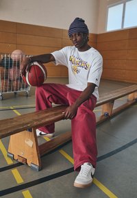 Bijela majica s ljubičastim i žutim natpisom "Lakers", crvene široke hlače, bijele i crne tenisice, sjedi na drvenoj klupi s košarkaškom loptom.