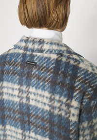 Nadkošile z vlněné směsi v modré, šedé a krémové kostkované vzoru s texturovaným povrchem. Obsahuje černou logo nášivku na zadním límci.