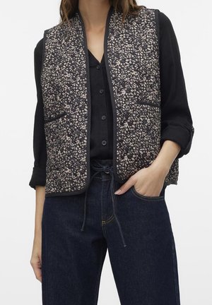 Femme portant une chemise noire à boutons avec manches retroussées, un gilet matelassé noir et beige à motif, et un jean bleu foncé avec une main dans la poche.