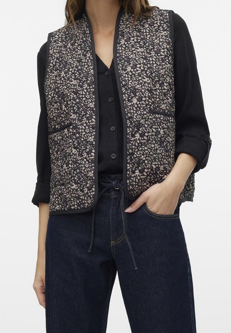 Femme portant une chemise noire à boutons avec manches retroussées, un gilet matelassé noir et beige à motif, et un jean bleu foncé avec une main dans la poche.