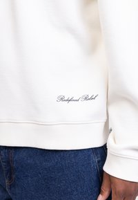 White sweatshirt made of textured fabric with ribbed cuffs and waistband. Features "Redefined Rebel" embroidered on the lower side.