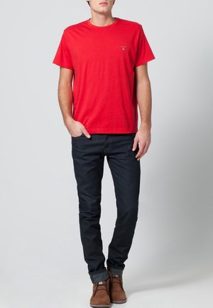 T-shirt en coton rouge avec un col rond, associé à un jean bleu foncé et des bottines brunes à la cheville. Design minimal, manches courtes et coupe ajustée.