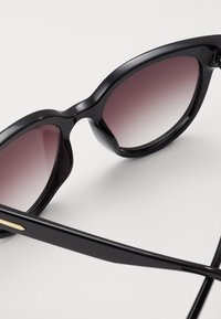 Black sunglasses with a rounded cat-eye shape, gradient lenses transitioning from dark to light. Sleek plastic frame with gold accent detailing.