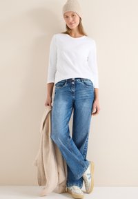 Chemise blanche à manches longues avec un col rond, jean bleu clair, bonnet en tricot beige, tenant une couche extérieure beige, et baskets beiges avec des accents bleus.