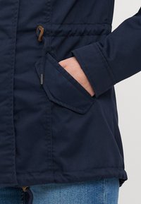 Veste marine avec une texture matelassée horizontale, poche latérale avec rabat et cordon de serrage à l'ourlet pour un ajustement réglable.