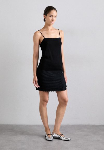 Black tank top with thin straps paired with a textured black skirt featuring a scalloped hem and button detail. Shiny silver flats complete the look.
