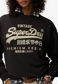 Zwarte sweatshirt met "SuperDry" logo in metallic goud, voorzien van geribbelde manchetten en een ruitkraag die eronder uitsteekt.