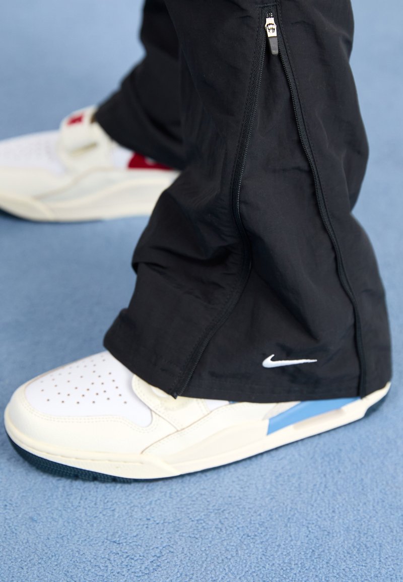 Nike-Sneaker mit weißem Obermaterial, durchlässiger Zehenpartie, blauen Akzenten, kombiniert mit schwarzen Hosen mit seitlichem Reißverschluss. Vor einem blauen Hintergrund.