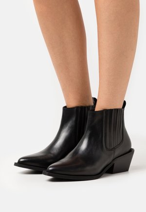 Chevilles et parties inférieures des jambes portant des bottines en cuir noir élégantes à bouts pointus, avec des panneaux élastiques sur les côtés et des talons bas inclinés, sur fond blanc.