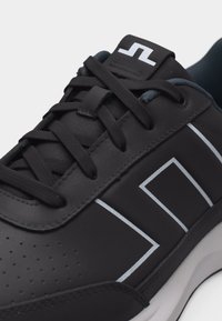 Μαύρο αθλητικό παπούτσι με μαύρα κορδόνια, λευκό γεωμετρικό σχέδιο στο πλάι, επένδυση στον αστράγαλο και μαλακή λευκή σόλα σε μονόχρωμο φόντο.