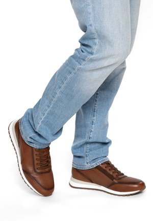 Person, die hellblaue Jeans und braune Ledersneaker mit weißen Sohlen trägt, steht mit einem Fuß vor dem anderen gekreuzt.