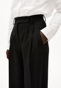 Zwarte geplooide broek met een hoge tailleband en zijzakken, gemaakt van een soepele stof. Gecombineerd met een witte overhemd met knopen.