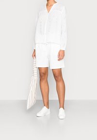Weiße, bestickte Bluse mit langen Ärmeln, kombiniert mit passenden Shorts. Die Accessoires umfassen eine gestreifte Tragetasche und weiße Sneakers.