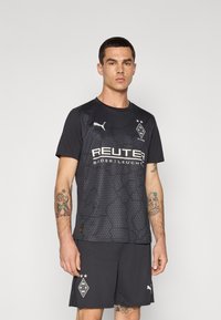 Svart sporttröja med korta ärmar, strukturerad design, med vitt tryckt logotyp och text. Kombinerad med svarta shorts och synliga tatueringar.