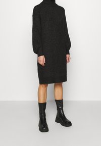 Abito nero lavorato a maglia con colletto alto, dotato di maniche lunghe e vestibilità ampia, abbinato a stivali neri in pelle alla caviglia.