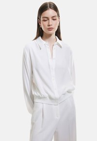 Camicia bianca con colletto, maniche lunghe e vita elasticizzata; texture liscia con una vestibilità rilassata, abbinata a pantaloni coordinati.