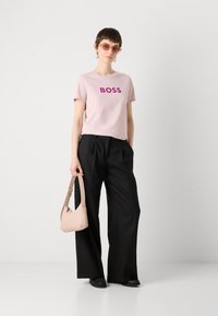 Rožinė medvilninė marškinė su užrašu „BOSS“ ryškiomis violetinėmis raidėmis, derinama su plačiomis juodomis kelnių ir šviesiai ruda petine rankine. Modelis dėvi akinius nuo saulės.