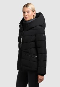 Geacă puffer neagră cu guler înalt, fermoar frontal și buzunare laterale. Are un design matlasat și o glugă detasabilă.
