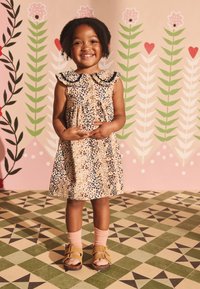 Kleopards printa kleita ar volāniem apkaklē, priekšā ar pogām, saskaņota ar rozā zeķēm un brūnām sandālēm uz rakstaina grīdas fona.