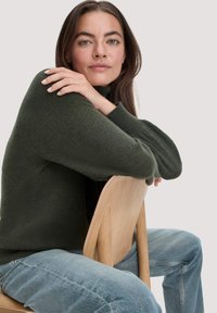 Dunkelgrüner Pullover mit hohem Kragen, grob gestrickter Textur und lockerer Passform, kombiniert mit hellblauen Jeans, die auf einem Holzstuhl sitzt.