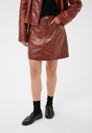 Personne portant une jupe en cuir marron et une veste assortie, un haut noir, des chaussettes noires et des mocassins noirs, debout avec une main dans la poche de la jupe.