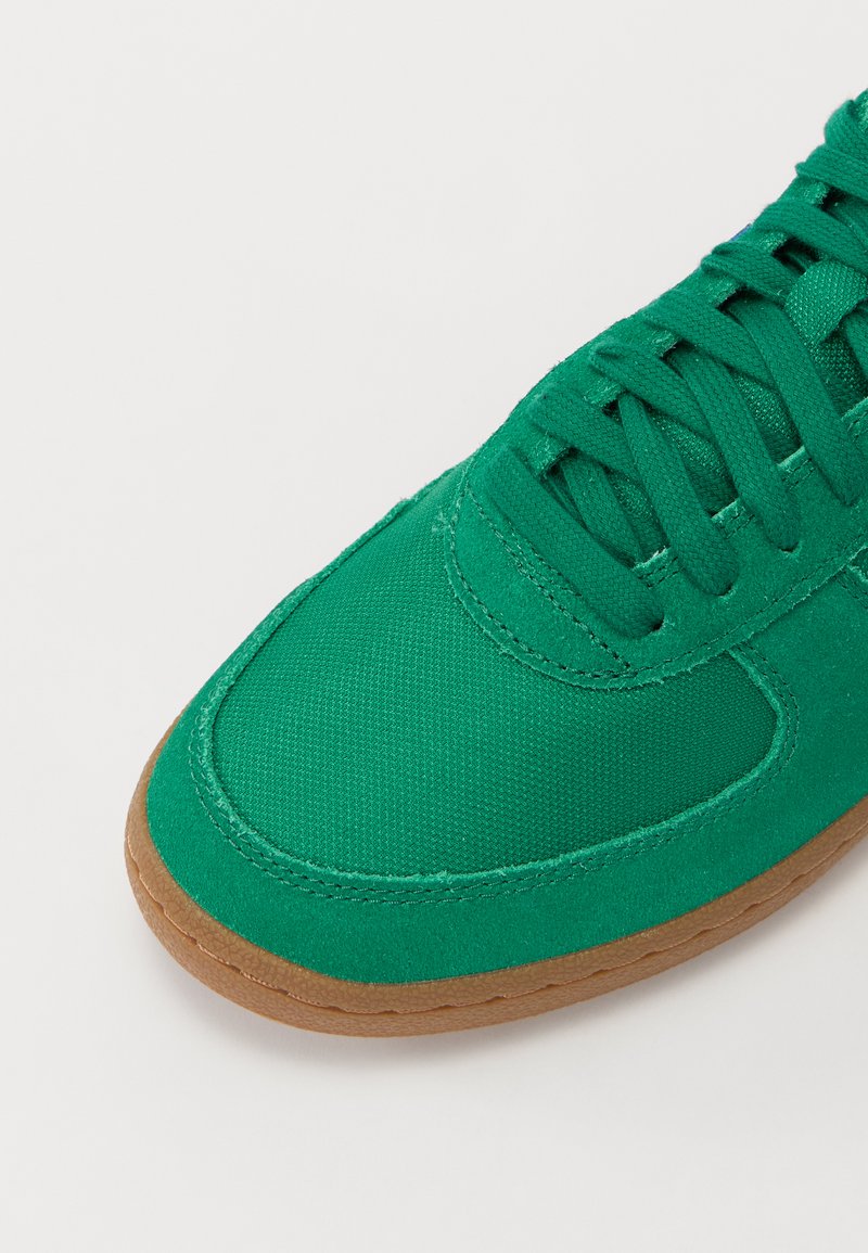 Sneaker in tela verde con punta arrotondata, superficie texturizzata, suola in gomma e design con lacci coordinati. Dettagli di cucitura evidenti.