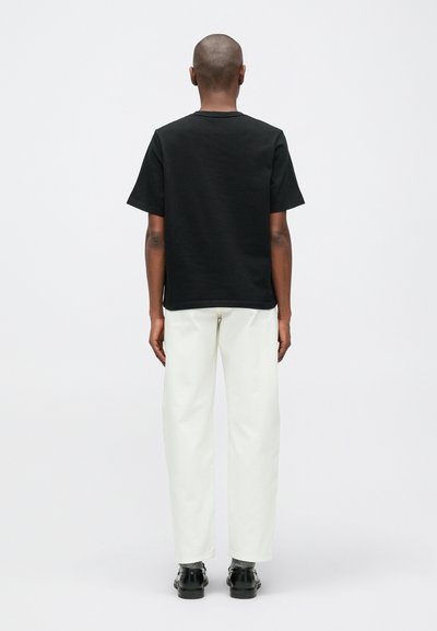 Osoba s oholenou hlavou, oblečená v černém tričku krátkého rukávu, bílých kalhotách, šedých ponožkách a černých botách, stojící zády k fotoaparátu na bílém pozadí.