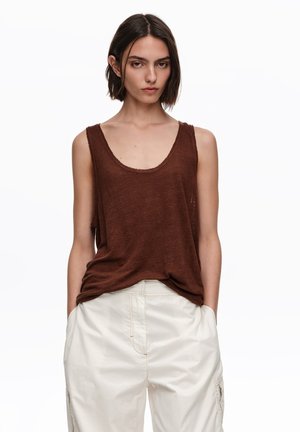 Mujer con cabello corto y castaño, lleva una blusa sin mangas marrón y pantalones blancos holgados, de pie con las manos en los bolsillos contra un fondo blanco.