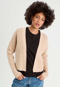 Cardigan beige à côtes avec un devant ouvert et des manches longues, porté sur un t-shirt noir. Texture remarquable avec une coupe décontractée et courte.