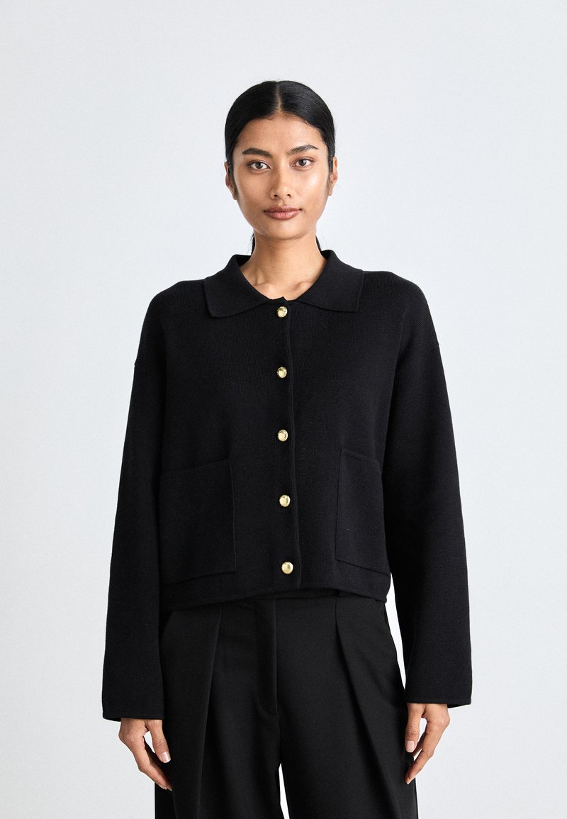 Black knitted jacket with a collar, featuring gold buttons and two front pockets. The design is cropped with long sleeves and a smooth texture.