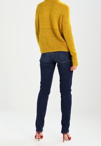 Suéter amarillo con cuello alto, combinado con jeans ajustados azul oscuro y zapatos de tacón rojos. Texturas suaves y diseño sencillo.