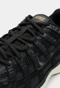 Černá atletická bota s prodyšným svrškem, plastovými akcenty a plochými tkaničkami. Výrazné logo Nike v zlaté barvě na jazyku. Texturovaný povrch.
