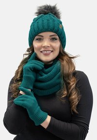 Gorro de punto en color verde azulado con un pompón de peluche sintético, guantes a juego en verde azulado y una bufanda gruesa. Presenta patrones acanalados y una textura suave.