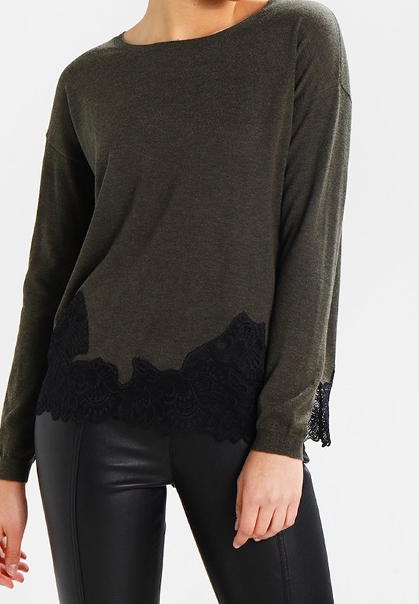 Femme portant un haut à manches longues vert olive avec une bordure en dentelle noire à l'ourlet, associé à un pantalon en cuir noir.