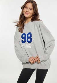 Szürke pulóver, laza szabással, kék hímzett "98" számmal és fehér "AHL. COLLEGE SPORTS" felirattal az elején. Puha tapintású, hosszú ujjú.