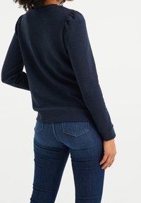 Marinblå texturerad pullovertröja med rund halsringning och puffärmar, parat med mörkblå tighta jeans. Diskret ribbad textur synlig.