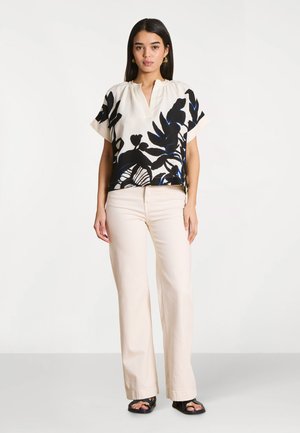 Femme portant un pantalon large crème, des sandales noires à enfiler et un chemisier blanc avec des motifs de feuilles noires et bleu foncé.