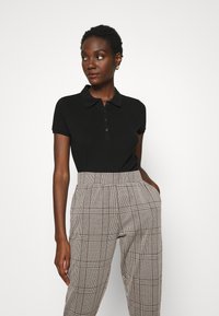 Camisa polo preta com colarinho clássico e placket de botões, combinada com calças de padrão xadrez de cintura alta em tons de bege e castanho.