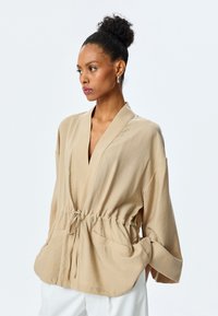 Beige leichte Jacke mit Wickeldesign, verstellbarem Taillenbund und zwei vorderen Taschen. Mit weiten Ärmeln und einer glatten Textur.