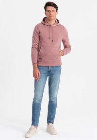 Felpa con cappuccio in cotone rosa polveroso con tasca frontale, abbinata a jeans azzurri e sneaker beige. Coulisse regolabili sul cappuccio.